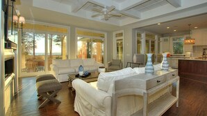 Un salon spacieux avec un grand canapé blanc, une table basse et une cheminée. On aperçoit un coin cuisine avec une table à manger et des chaises à travers une porte ouverte.