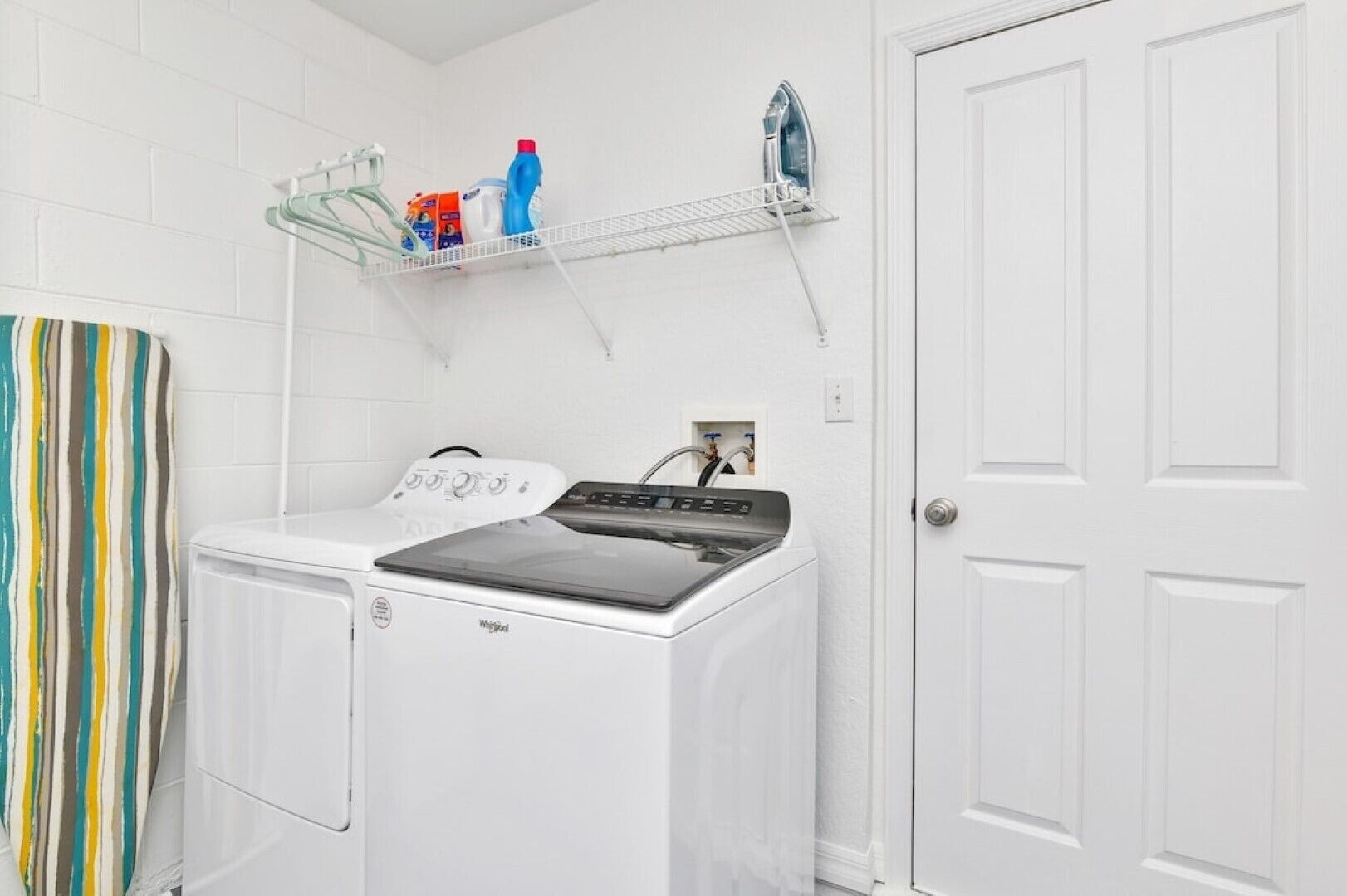 A laundry area with a front-loading washer and dryer, a shelf with cleaning supplies, and a striped shower curtain.