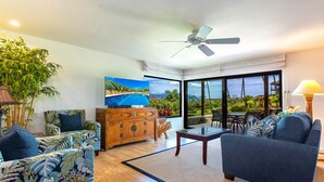 A spacious living room with a large window, a ceiling fan, a wooden cabinet, and a sofa. - Elua 2303: Gorgeous Luxe Condo, Ocean Sunset Views (Kihei)