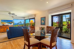 A dining area with a glass table and wooden chairs, a blue sofa, and a view of a pool and tropical plants.
