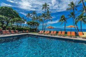 Ein Swimmingpool mit Liegestühlen und Sonnenschirmen, umgeben von Palmen und Blick auf den Ozean.