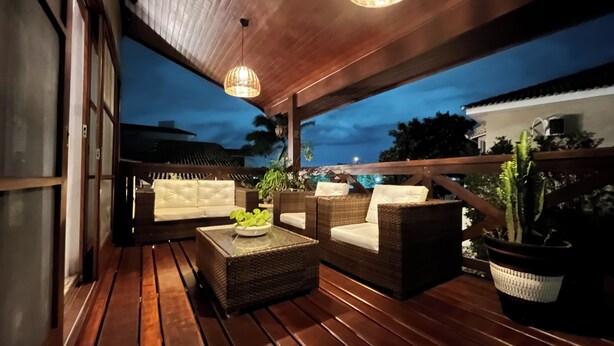 Un espacio de estar al aire libre con mobiliario de mimbre y una terraza de madera, con vista a una zona residencial al anochecer.