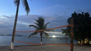 Una red de voleibol de playa con un poste de madera y una malla naranja, palmeras y vistas al mar y luces de la ciudad a lo lejos.