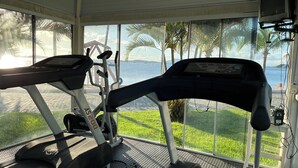 Una caminadora con pantalla digital, ubicada en una habitación con ventanas amplias que dan a un cuerpo de agua y palmeras.