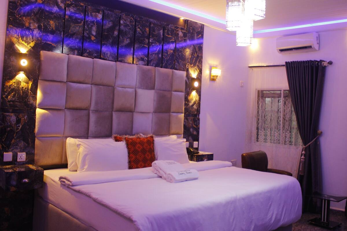 A hotel room with a large bed, a headboard, a nightstand, a desk, a chair, a window with curtains, and an air conditioning unit.