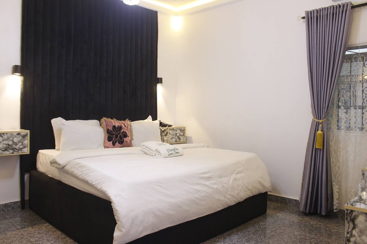 A neatly made bed with white bedding, a dark headboard, and decorative pillows. There are two bedside tables with lamps and a window with curtains.