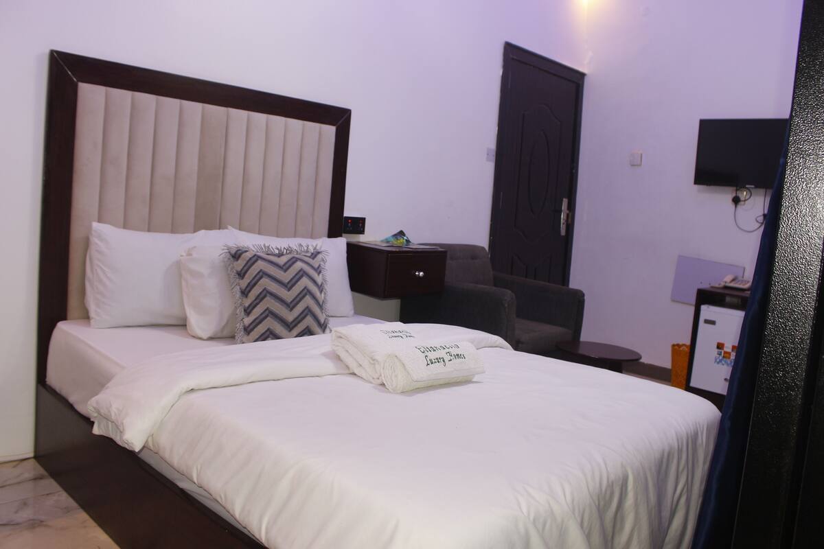 A neatly made bed with a folded towel, a bedside table, a chair, a TV, and a door in a hotel room.