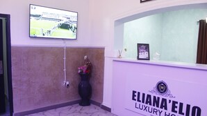 Ein Empfangsbereich mit an der Wand montiertem Fernseher, der ein Sportevent zeigt, einem Empfangstresen mit der Aufschrift "ELIANA'ELIO LUXURY HOMES" und einer auf dem Boden stehenden Vase mit Blumen.