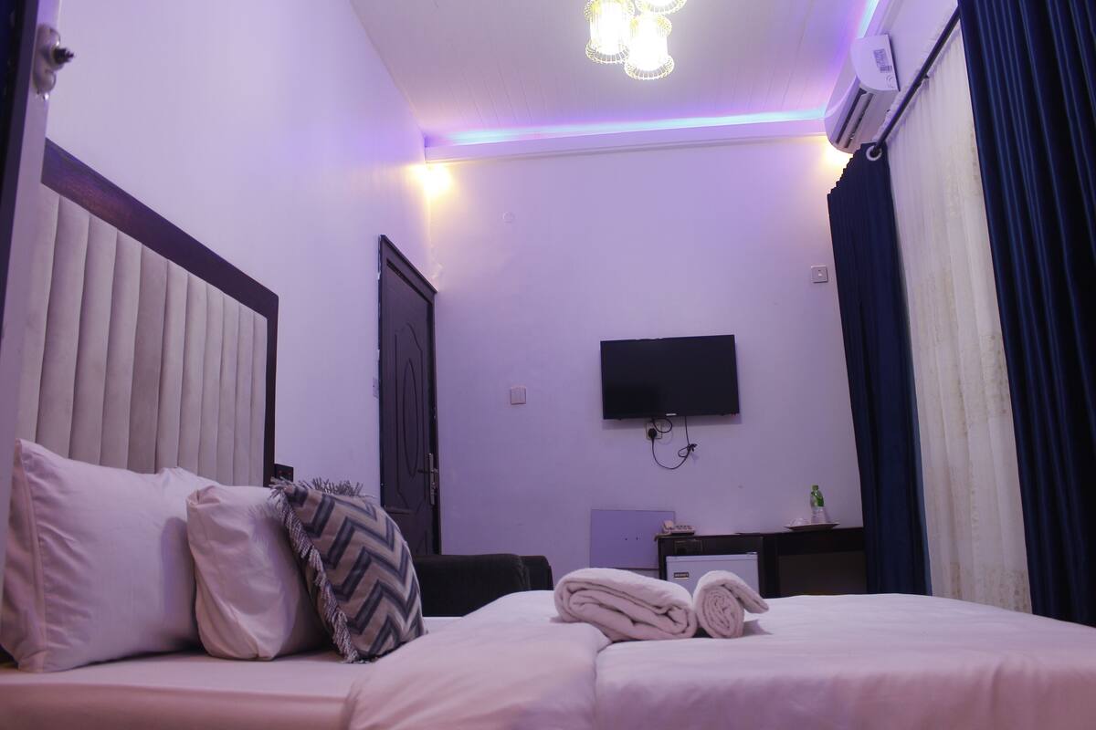 A hotel room with a bed, pillows, a flat-screen TV, a desk, and a window with curtains.
