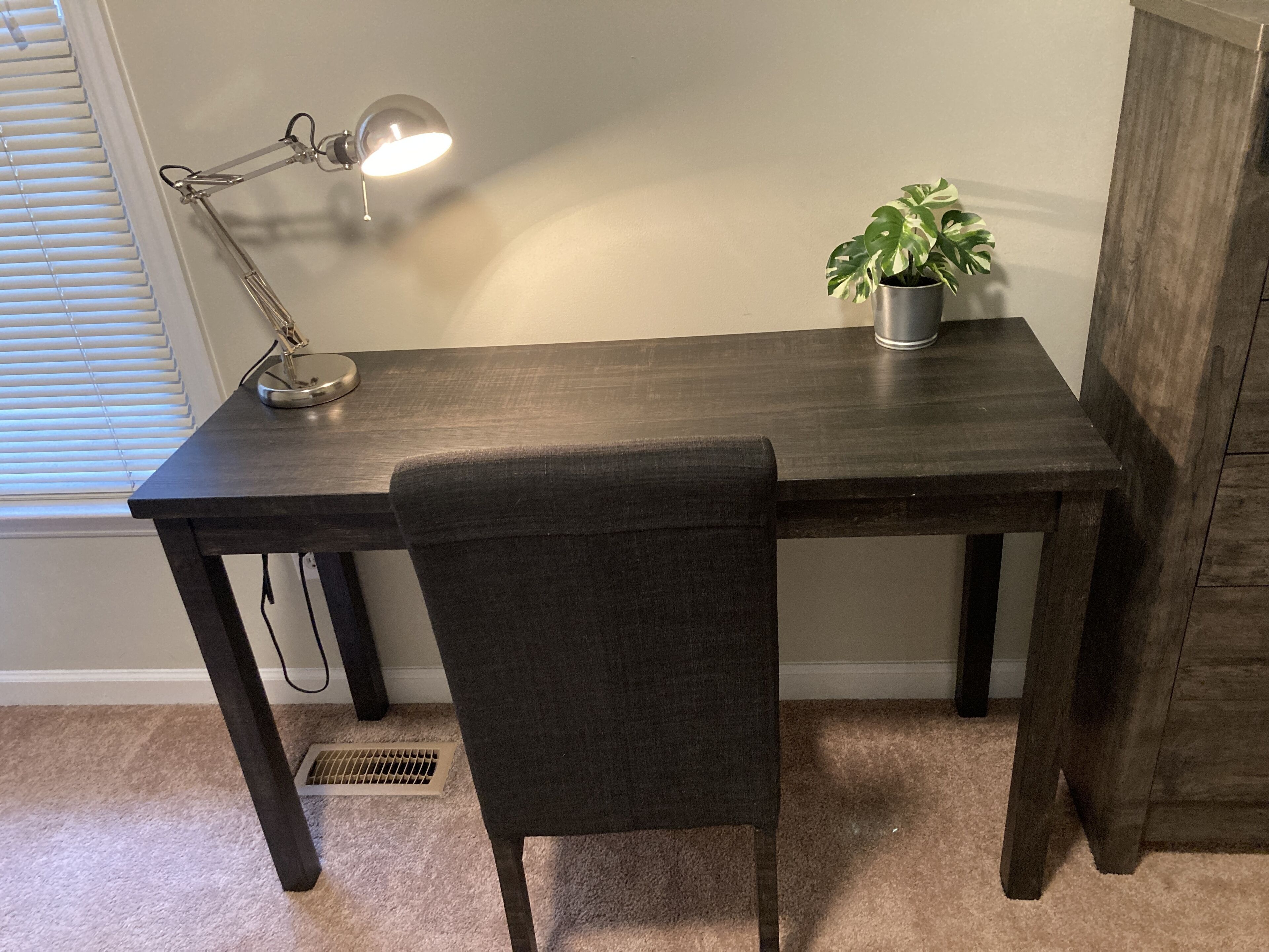 Un escritorio de madera oscura con una lámpara, una planta en maceta y una silla en una habitación con alfombra color crema y una ventana con persianas.