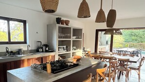 A modern kitchen with a central island, wooden chairs, hanging woven light fixtures, and a view of the outdoors.