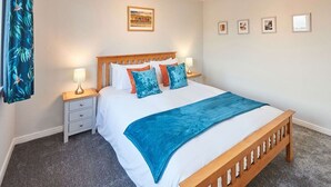 Ein ordentlich eingerichtetes Schlafzimmer mit einem hölzernen Bett, weißer Bettwäsche und einer blauen Tagesdecke. Es gibt zwei Nachttische mit Lampen, ein an der Wand hängendes Bild und einen Vorhang mit Blätermotiv.
