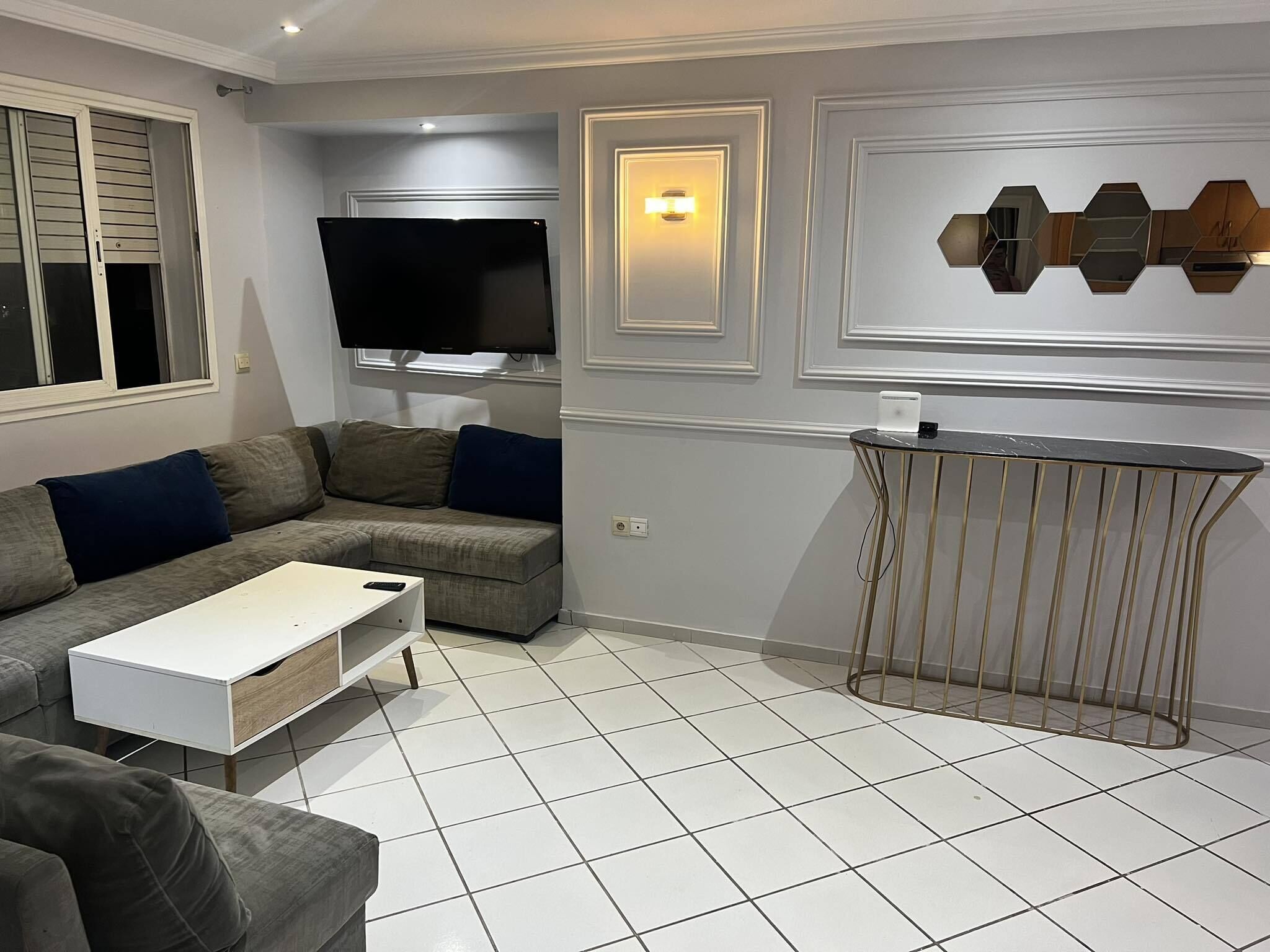 A modern living room with a grey sofa, a white coffee table, a TV mounted on the wall, and a decorative wall piece.