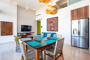 Een moderne keuken met een roestvrijstalen koelkast, een eethoek met een houten tafel en rieten stoelen, en een aan de muur bevestigde televisie.