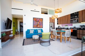 Un salon moderne avec un canapé bleu, un coin repas comprenant une table et des chaises, et une cuisine dotée de meubles en bois et d’appareils électroménagers.