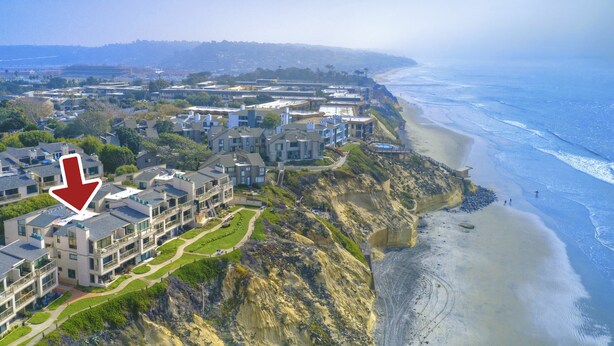 An aerial view of a coastal residential area with modern buildings, a beach, and a cliff.