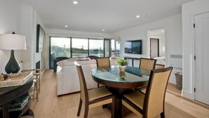 A modern living room with a glass dining table, white sofas, and a large window offering a city view.