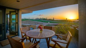 An outdoor patio with a round wooden table and chairs, a vase with flowers, and a view of the sunset over a landscaped area.