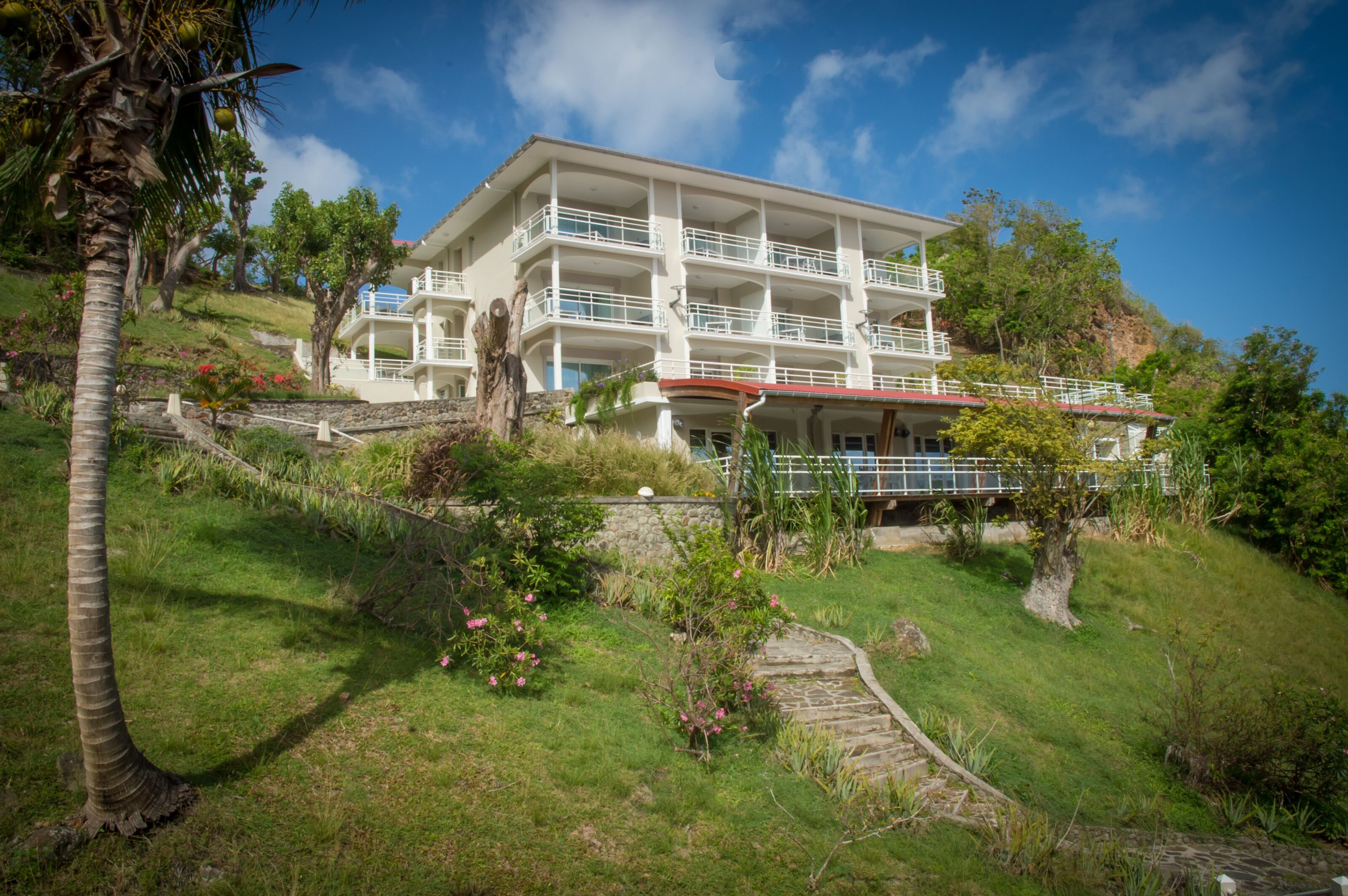 Un hôtel à plusieurs étages doté de balcons, situé sur une colline verdoyante, agrémentée de palmiers et de plantes à fleurs.