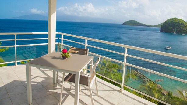 Un balcon avec une table et des chaises, donnant sur une mer d’un bleu limpide, avec un petit bateau et une île qui se dessinent à l’horizon.