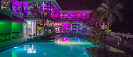 A pool area with a bridge, palm trees, and a building with colorful lighting.