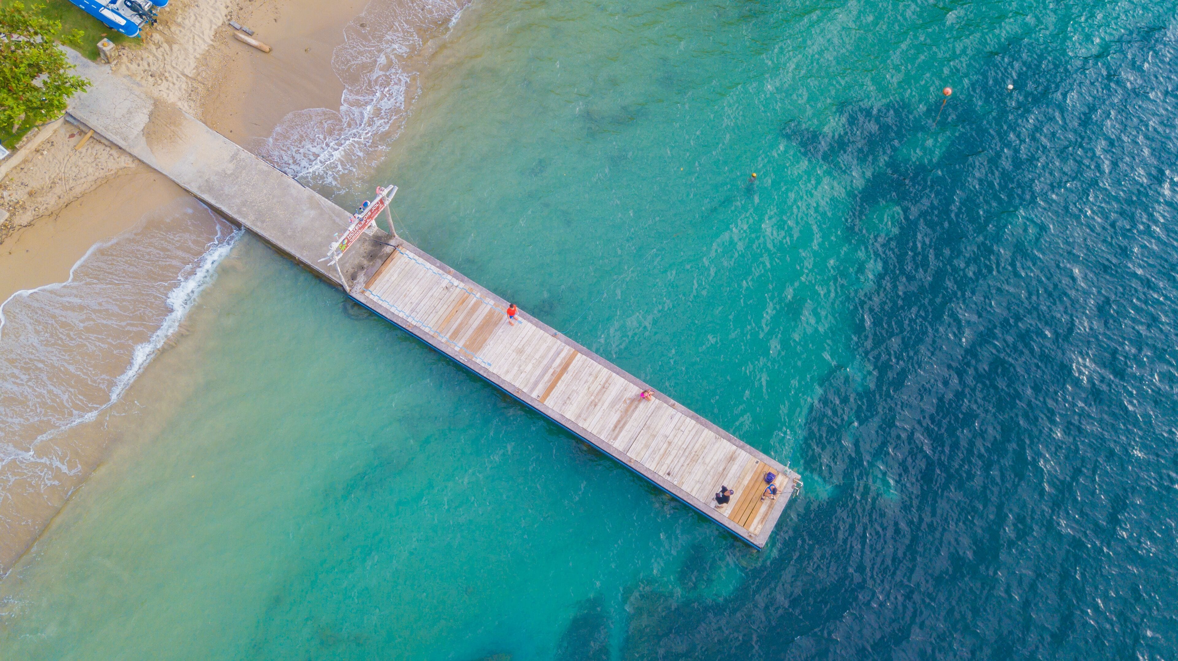 Vista aérea de un muelle que se adentra en aguas turquesas, con una playa de arena a un lado.