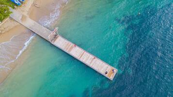 Vista aérea de un muelle que se adentra en aguas turquesas, con una playa de arena a un lado.