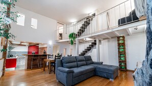 Ein geräumiger Wohnbereich mit einem großen, grauen Ecksofa, einem Holzespeisetisch mit Stühlen, einer Küche mit roten Akzenten und einer Treppe, die zu einer oberen Ebene führt.