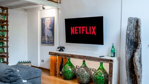 Ein Wohnzimmer mit einem Fernseher, der "NETFLIX" zeigt, einer Couch, einem Holztisch mit grünen Vasen und einem Holzstuhl.