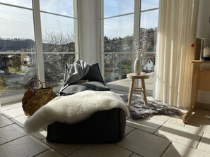 Quarto moderno com uma grande janela, uma cama aconchegante com manta de pele, um pequeno banquinho de madeira e um vaso com flores.