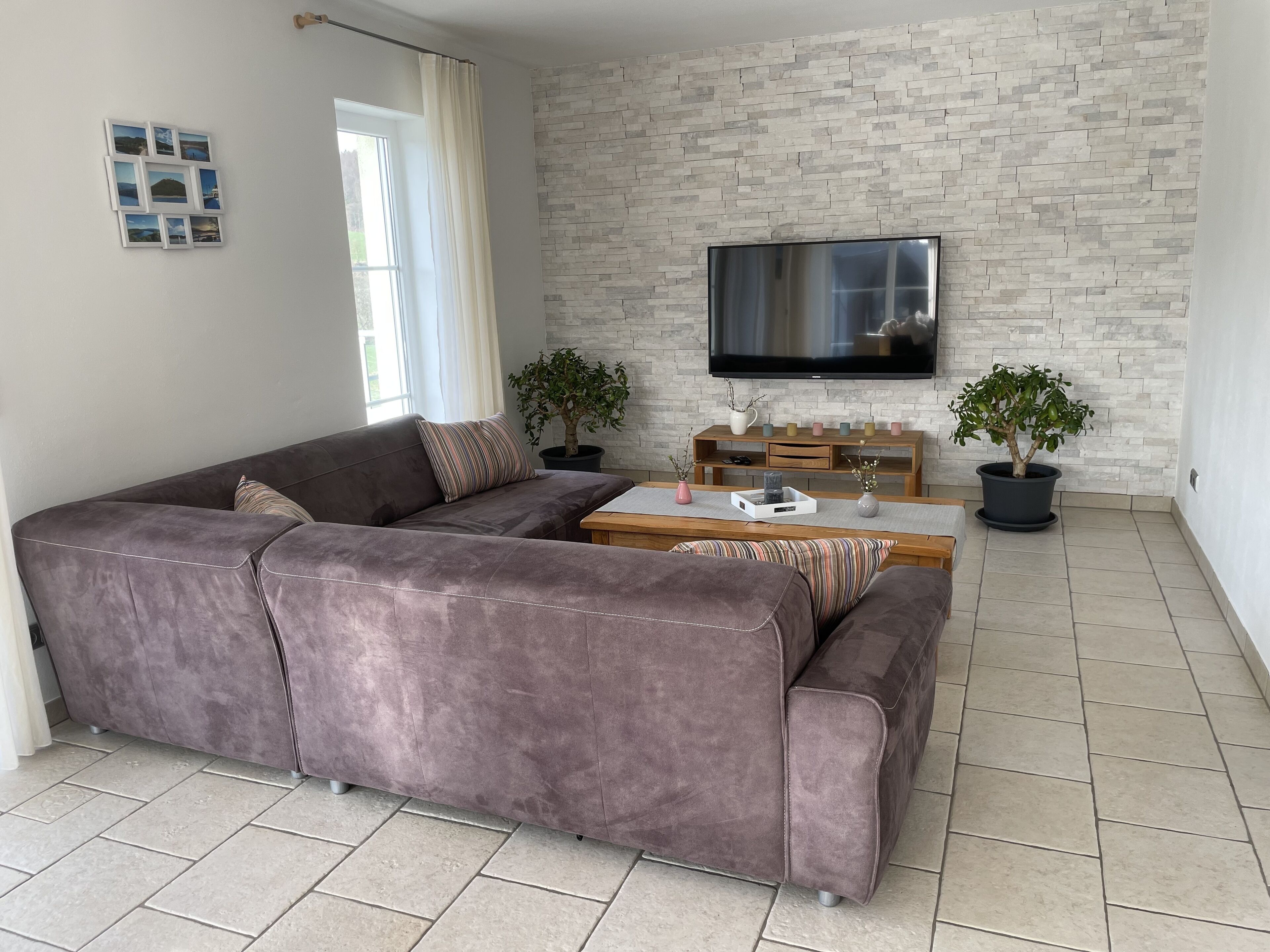 Una sala de estar con un sofá seccional grande, un televisor de pantalla plana montado en una pared de piedra, una mesa de centro y dos plantas en macetas.