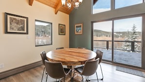 A dining area with a round wooden table, four chairs, and a chandelier. There are framed pictures on the walls and large windows offering a view of a snowy landscape. - The Fishermen’s Place: Mountain Views + Dog Friendly (Breckenridge)