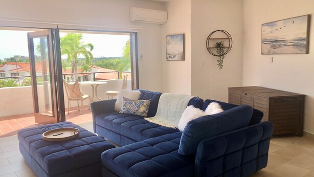 Un salon avec un canapé bleu, un pouf et une petite table sur laquelle se trouve un plateau. Il y a un balcon d’où l’on peut admirer des palmiers et des maisons.