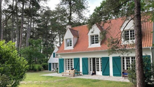 Une maison à deux étages, avec un toit recouvert de tuiles rouges et des murs blancs, entourée d’une cour verdoyante et d’arbres.