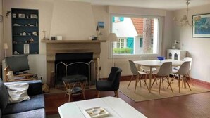 Un salon avec une cheminée, un coin repas avec une table et des chaises, une bibliothèque et une fenêtre donnant sur une maison.