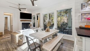Un comedor con una mesa de madera, bancos y sillas. Tiene un ventilador de techo, una chimenea y ventanas grandes con vistas al exterior.