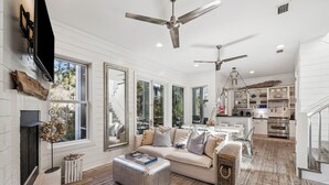 Un salón amplio con un sofá grande, un reposapiés y una chimenea. Se ven varias ventanas y una zona de cocina al fondo.