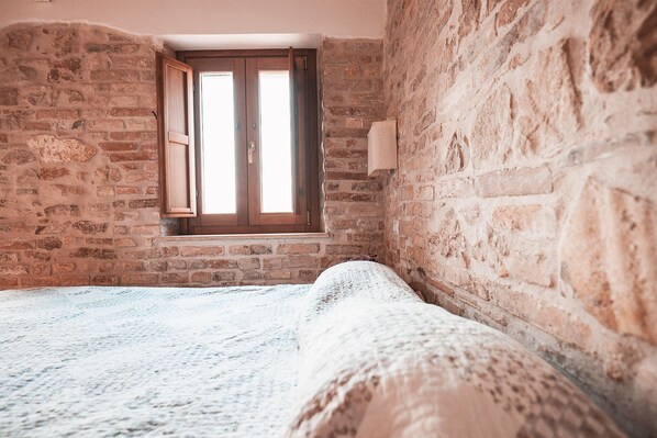 Una habitación con paredes de ladrillo visto, una ventana de madera y una cama con colcha blanca.