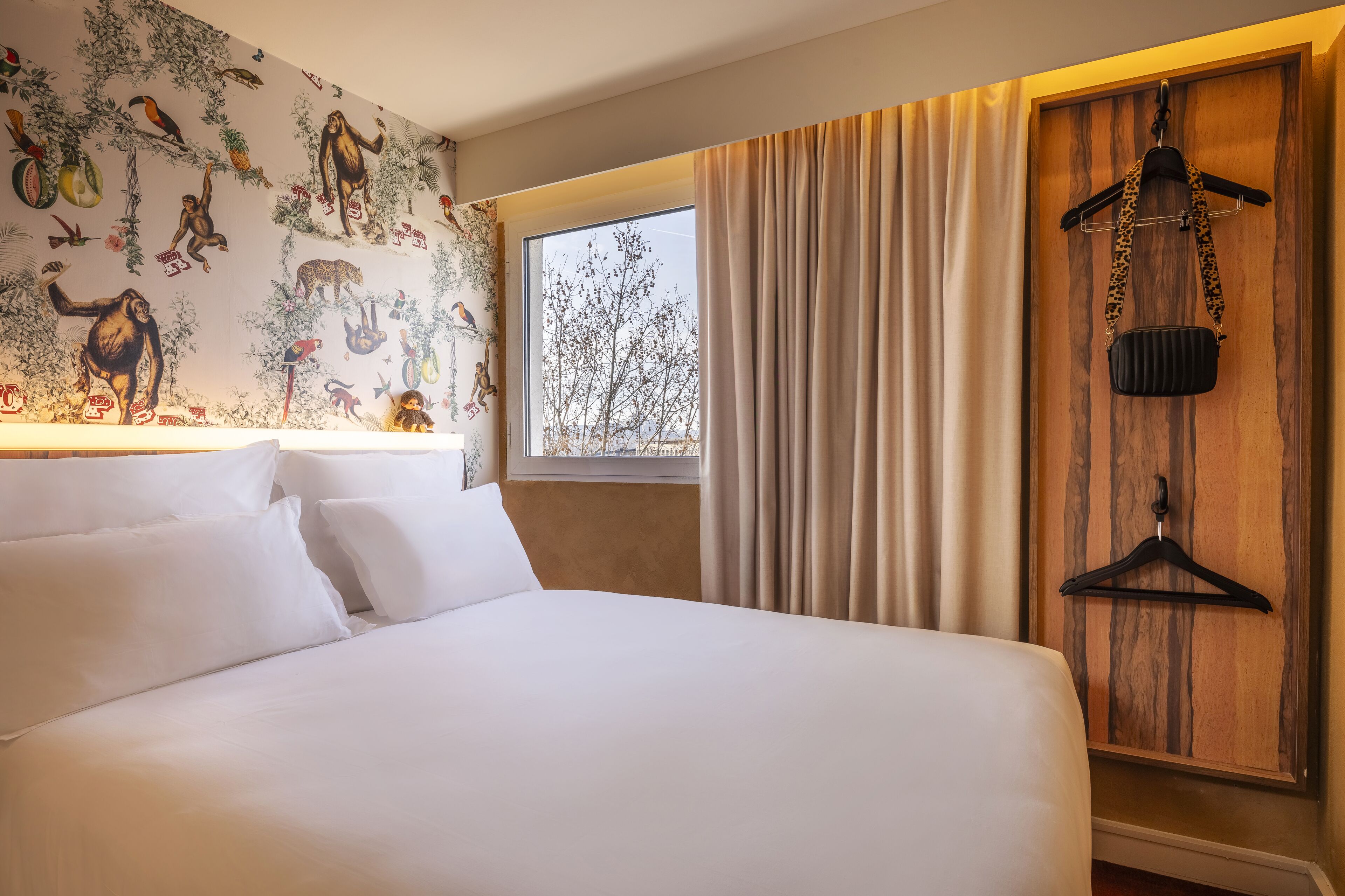 A hotel room with a large bed, a window with curtains, a wall with a mural, and a coat rack with a handbag.