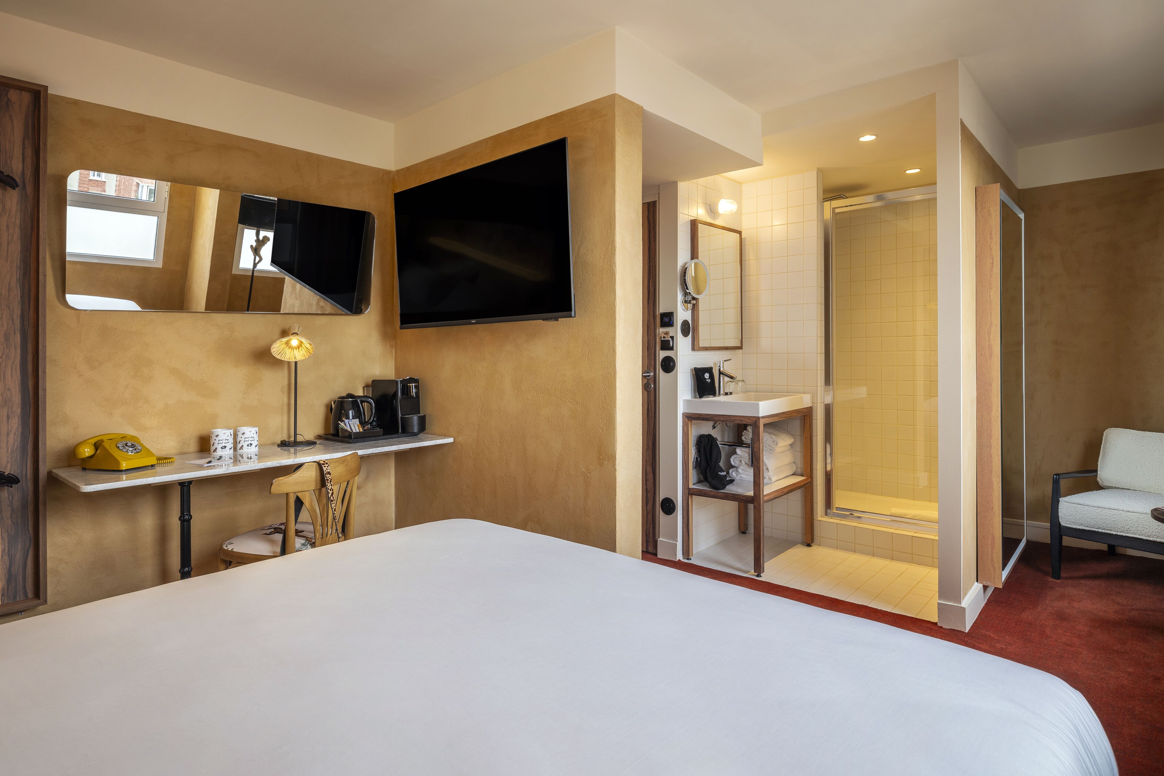 Une chambre d’hôtel avec un grand lit, un téléviseur fixé au mur, un bureau avec une lampe, un téléphone et un espace douche équipé d’une chaise.