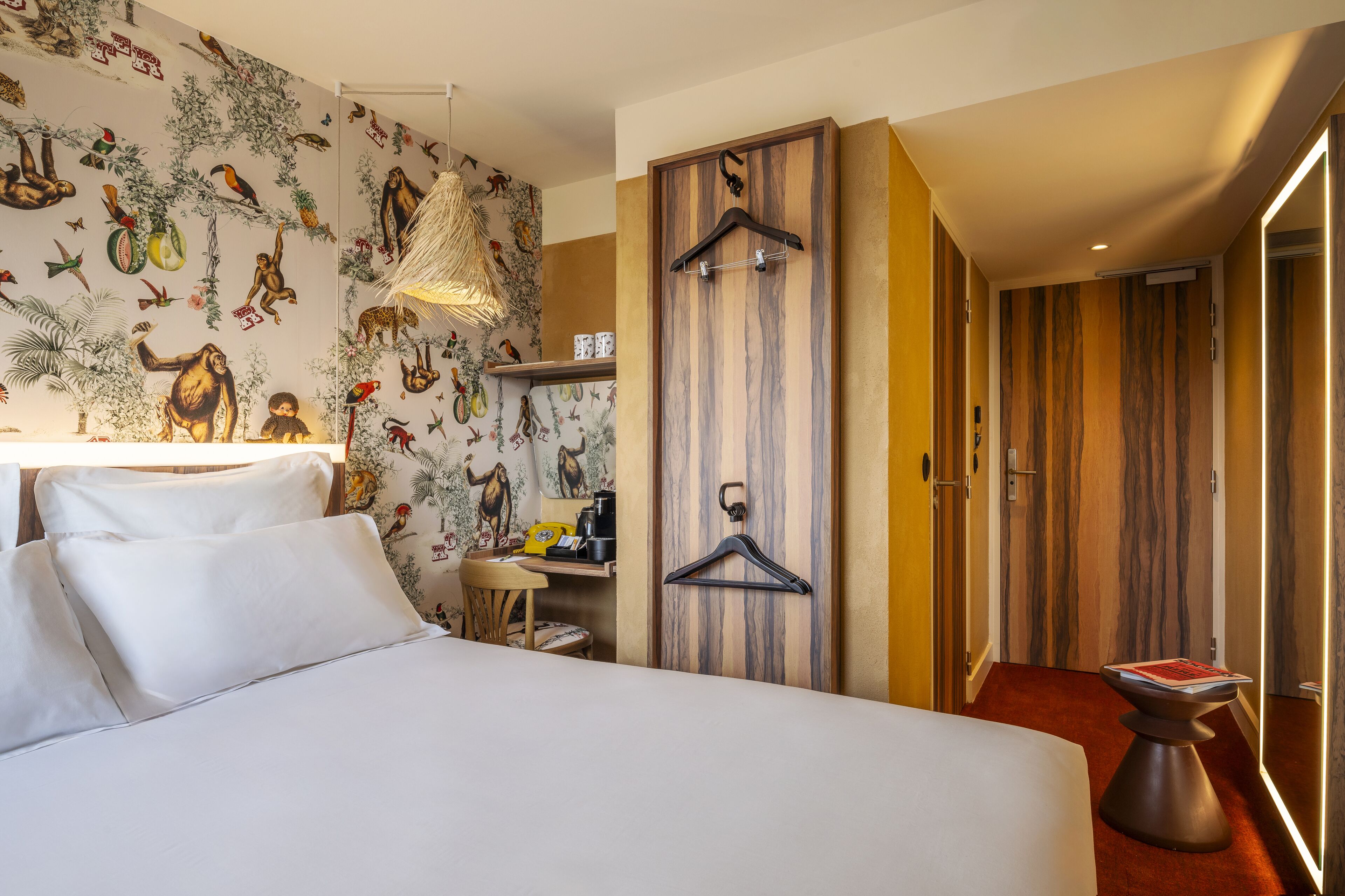 A hotel room with a bed, a desk with a chair, a hanging light fixture, and a wardrobe with a wooden door.