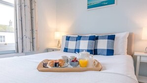 Ein ordentlich bezogenes Bett mit einem karierten Kissen, ein Tablett mit Croissants, Obst und Orangensaft sowie eine Lampe auf einem Nachttisch.