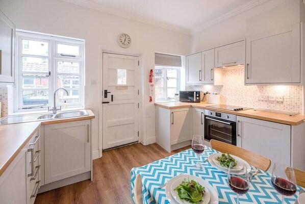 A compact kitchen with white cabinets, a microwave, and a dining area with a blue and white tablecloth. - Pelican Cottage, Aldeburgh (Aldeburgh)