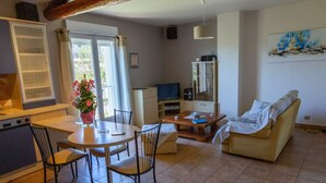 A living room with a dining table, chairs, a sofa, a TV, and a kitchen area with a refrigerator and a window with curtains. - L'Oustau de Famiho Air-conditioned gite with pool in renovated farmhouse 10 km from Avignon (Châteauneuf-de-Gadagne)