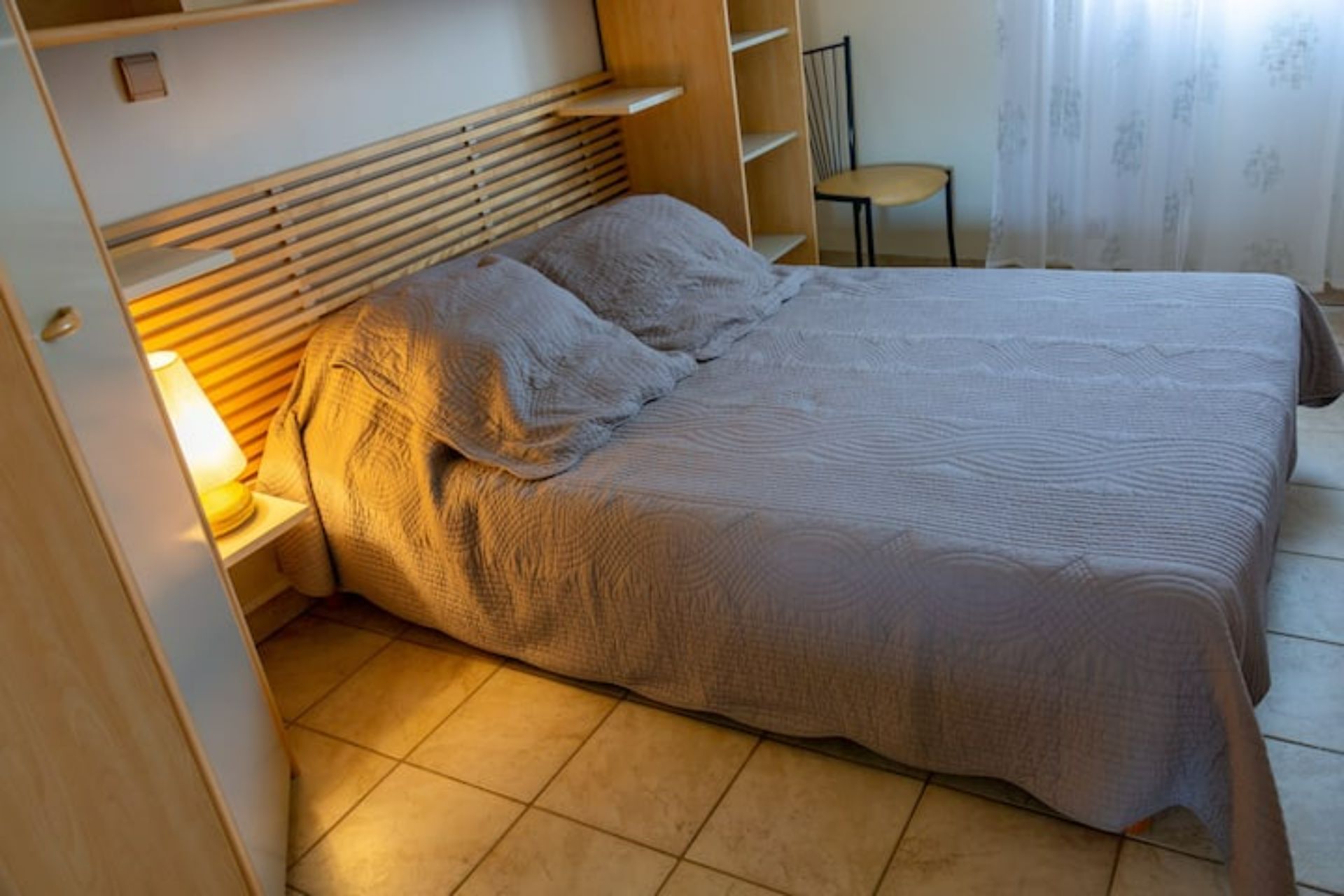 A neatly made bed with a headboard, a bedside lamp, and a chair in a room with a tiled floor and a window with curtains.