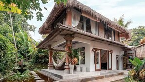 Ein traditionelles Haus mit einem Reetdach und einer Veranda, umgeben von üppiger Vegetation und einem kleinen Wasserspiel.
