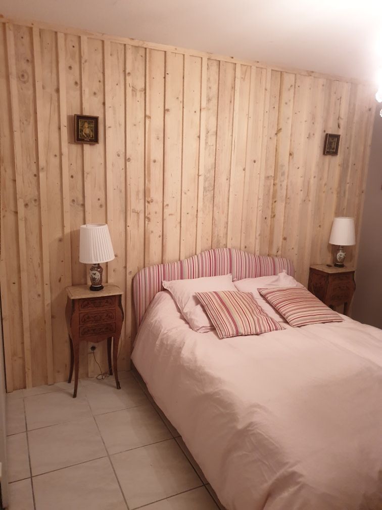 A bedroom with a wooden panel wall, a bed with striped bedding, two bedside tables with lamps, and a small drawer unit.
