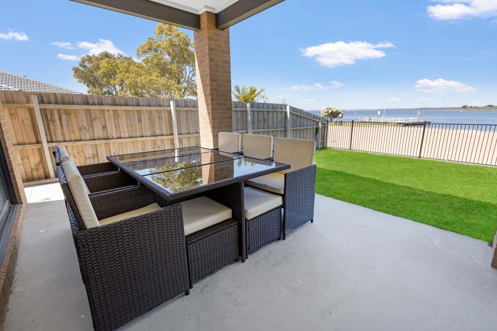 Une terrasse extérieure aménagée avec des meubles en osier et une table à plateau de verre, offrant une vue sur une pelouse et une étendue d’eau.