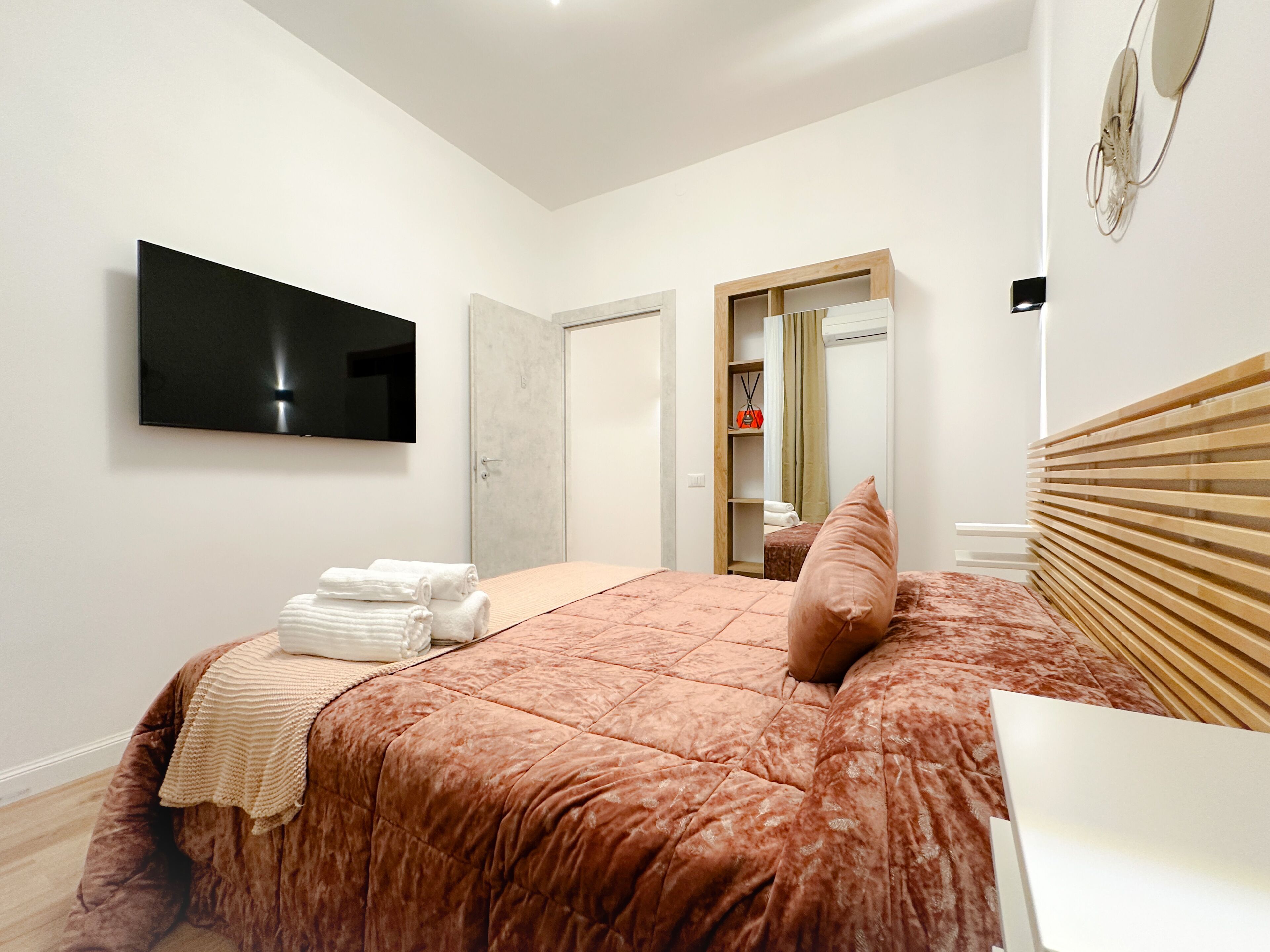 A bedroom with a bed, a TV mounted on the wall, a wooden headboard, and a mirror.