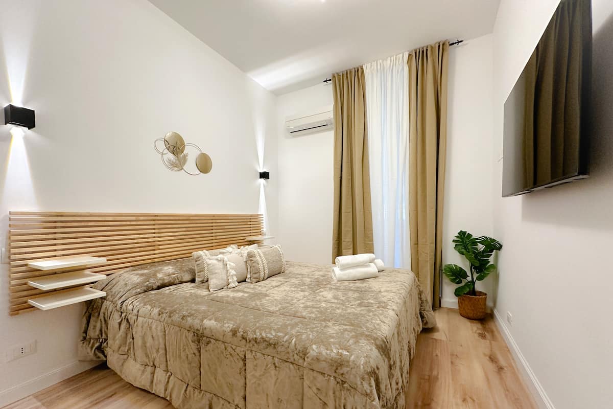 A modern bedroom with a wooden headboard, a marble-patterned bedspread, a potted plant, and a mounted TV.